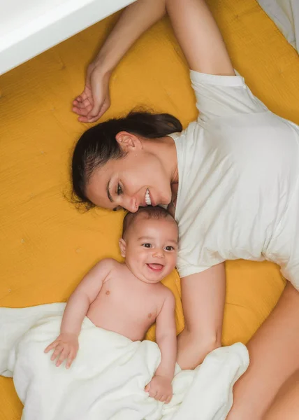 D'en haut de joyeuse mère aux cheveux noirs gaie en tenue décontractée couchée sur le lit avec contenu petit bébé en couverture — Photo de stock