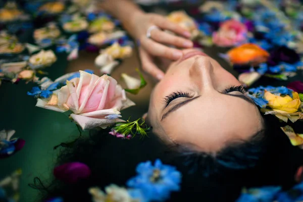 Desde arriba atractiva cara femenina tocando mientras está acostado en la bañera con agua tibia y varias flores de colores - foto de stock