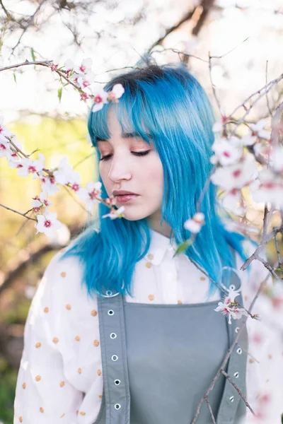 Joven mujer elegante con el pelo largo y azul con los ojos cerrados usando la fragancia general de moda disfrutando de la floración del árbol mientras está de pie en el jardín de primavera - foto de stock