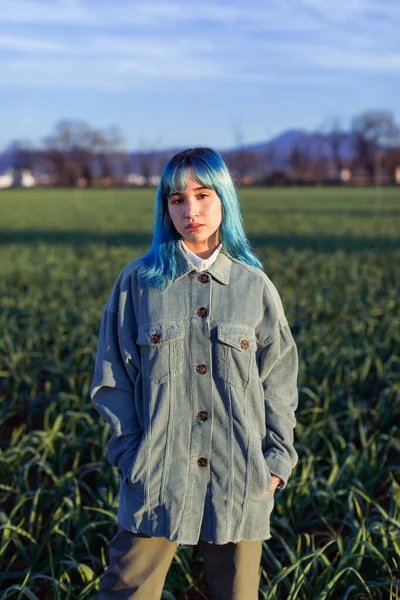 Thoughtful young female with blue hair looking at camera dressed in trendy jacket standing in green field in sunny evening — Stock Photo