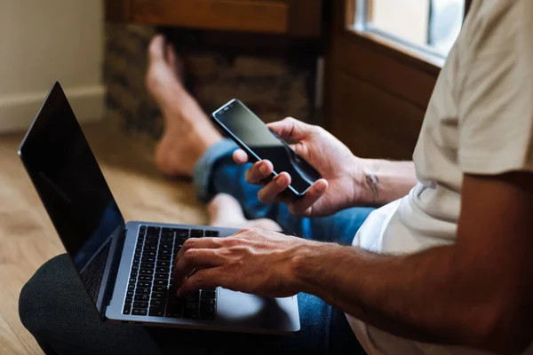 Hombre irreconocible sentado en el suelo y usando el ordenador portátil y el teléfono inteligente mientras trabaja en un proyecto remoto en casa - foto de stock