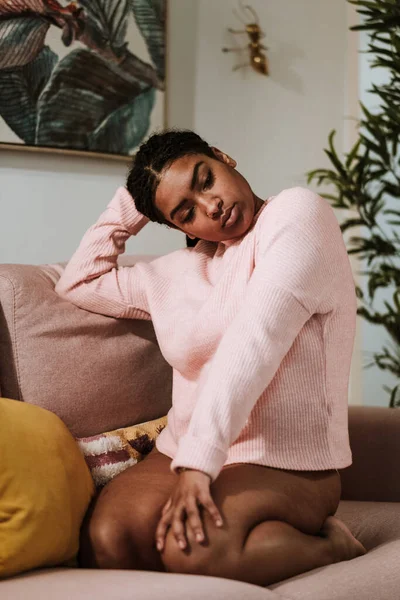Nachdenklich gelangweilte junge Afroamerikanerin in Freizeitkleidung sitzt auf dem Sofa, während sie den Tag zu Hause verbringt — Stockfoto