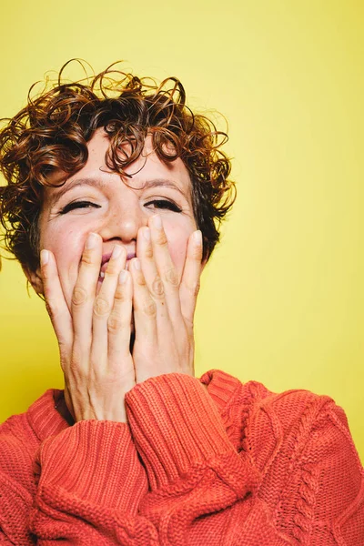 Erstaunlich lachende Frau mit lockigem Haar trägt orangefarbenen Strickpullover, der Mund mit Händen bedeckt und wegschaut, während sie vor gelbem Hintergrund steht — Stockfoto