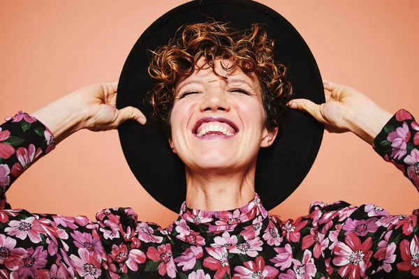 Optimistic young beautiful female model with curly hair wearing stylish black hat and colorful blouse with closed eyes smiling while standing against orange background — Stock Photo