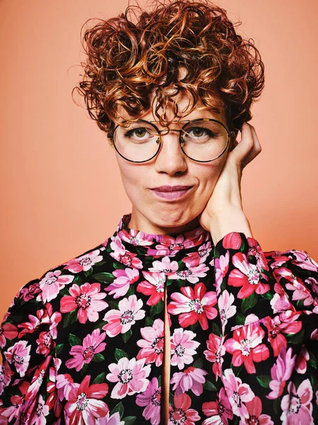 Nachdenklich zweifelhaft schöne lockige Frau in trendiger Brille und stilvoller bunter Bluse mit floralem Ornament blickt in die Kamera vor rosa Hintergrund — Stockfoto