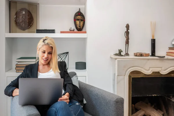 Cheerful blonde businesswoman smiling and browsing laptop while sitting in comfortable armchair near fireplace in cozy room working remotely from home — Stock Photo