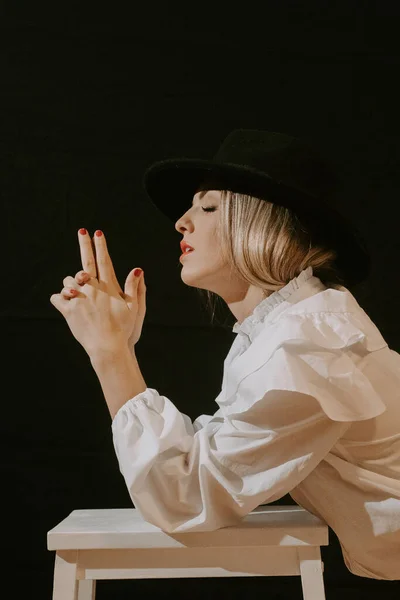 Side view of young blonde woman in white blouse and stylish hat with closed eyes while leaning on stool making gun gesture with fingers against black background — Stock Photo