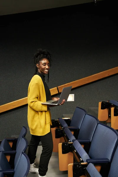 Ganzkörper-Afroamerikanerin in lässiger Kleidung blickt in die Kamera und blättert im Laptop, während sie inmitten von Sitzreihen im modernen Hörsaal steht — Stockfoto