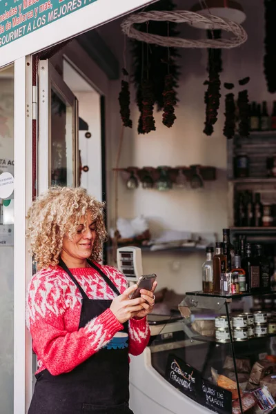 Positive adult woman using mobile phone while standing near entrance of local delicatessen food store — Stock Photo