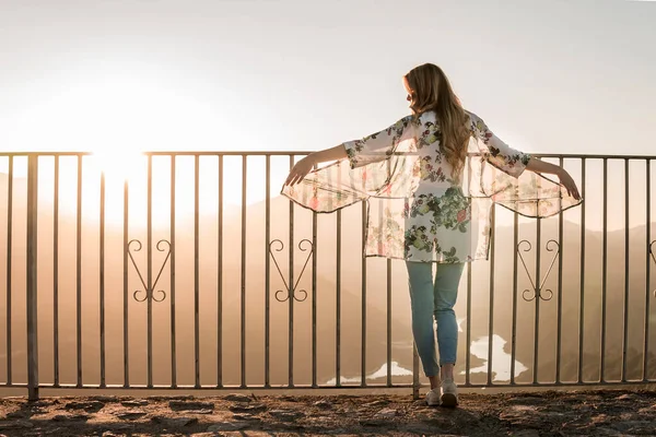 Vista trasera de la turista femenina en ropa casual de pie cerca de la valla de metal y admirando la vista del atardecer durante el paseo en el día de verano - foto de stock