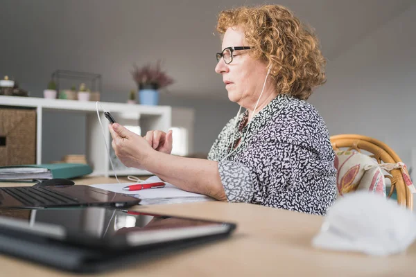 Visão lateral da mulher adulta focada em roupas casuais e óculos usando smartphone em fones de ouvido enquanto se senta à mesa e trabalha em laptop em casa — Fotografia de Stock