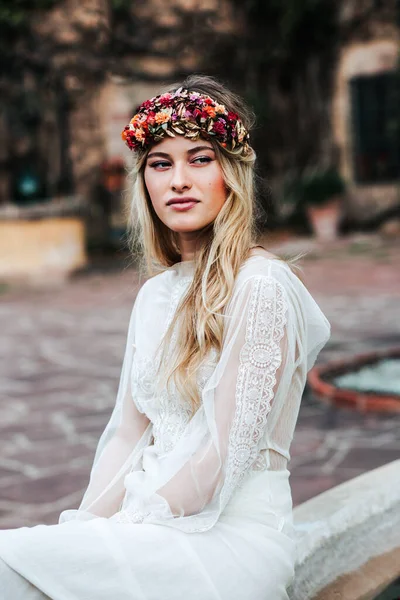 Pretty young woman in translucent white dress and floral wreath looking away while sitting on blurred background of yard on wedding day — Stock Photo