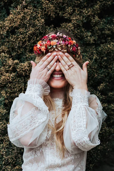 Delighted young female in bridal dress and floral wreath smiling and covering eyes while standing near green shrub in garden — Stock Photo