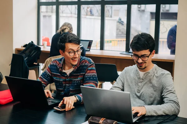 Позитивні співробітники у повсякденному одязі та окулярах сидять за столом і працюють над проектами по ноутбуках під час роботи в сучасному офісі. — стокове фото