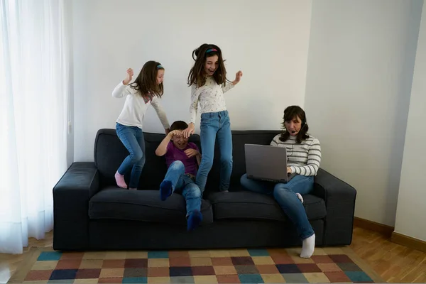 Group of positive smiling kids in casual jeans and shirts laughing and jumping on sofa while their mother working on laptop with headset nearby — Stock Photo