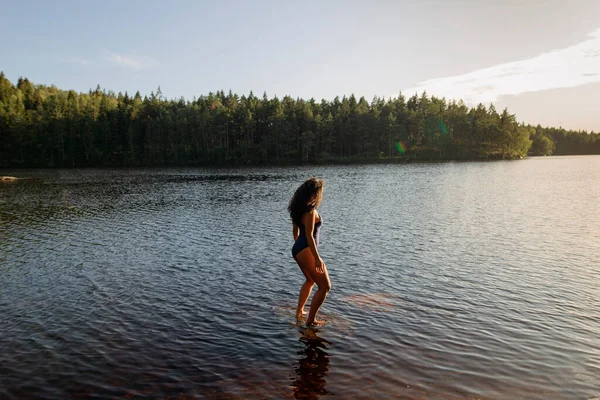 Desde arriba vista lateral de hembra delgada en traje de baño de pie en aguas tranquilas del lago mientras disfruta de la puesta del sol y el paisaje majestuoso - foto de stock
