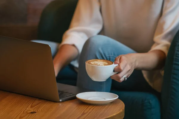 Ritagliato irriconoscibile giovane donna freelance in abiti casual con gustosa bevanda calda mentre seduto al tavolo rotondo in legno e utilizzando netbook in luce caffè moderno — Foto stock