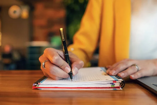 Crop donna d'affari irriconoscibile in eleganti vestiti colorati scrittura con penna nel taccuino mentre seduto al tavolo di legno e utilizzando il computer portatile in luce ufficio contemporaneo — Foto stock