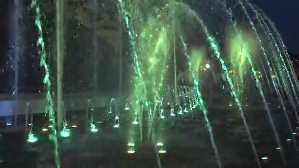 Fountain in night-time downtown. The water then rises up again it falls. At the bottom of the fountain lights shine in different colors. — Stock Video