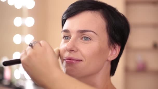 La chica sonríe mientras el maquillador aplica polvo con un pincel grande. En el contexto del lugar de trabajo iluminado . — Vídeo de stock
