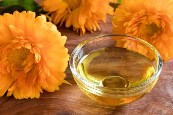 Calendula oil in a glass bowl with calendula flowers in the back — Stock Photo, Image