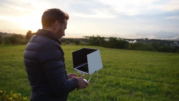 Un hombre barbudo vestido con la chaqueta baja mirando su tableta y sonriendo mientras pilotaba el dron en los campos, enfoque selectivo, en la puesta del sol — Vídeos de Stock