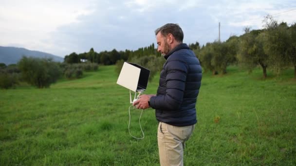 En skäggig man iklädd dunjacka titta på sin surfplatta och ler medan lotsa drönaren i fälten selektivt fokus, på solnedgången — Stockvideo