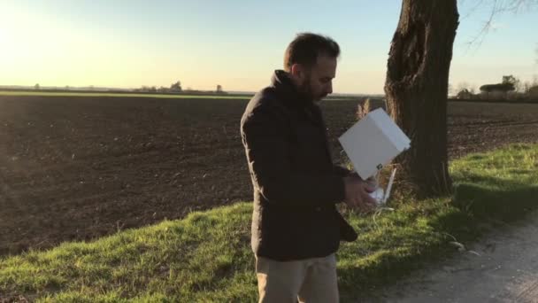 Ein bärtiger Mann in Daunenjacke steuert seine Drohne auf der Landstraße, bei Sonnenuntergang — Stockvideo