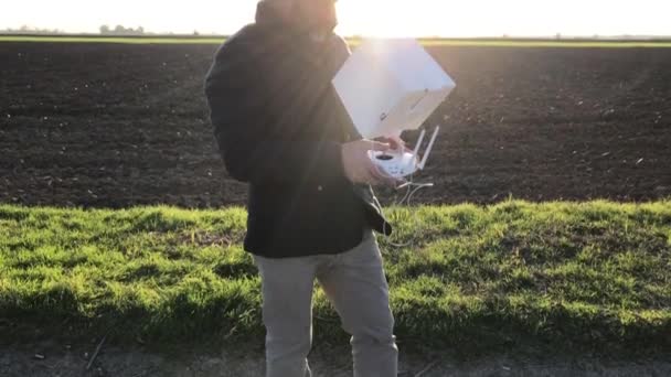 Um homem barbudo vestido de jaqueta pilota seu drone na estrada do campo, ao pôr do sol — Vídeo de Stock