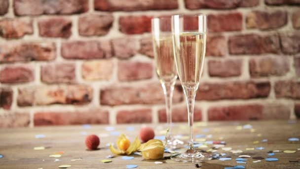 Vidéo au ralenti de beaucoup de confettis à la suite de deux flûtes de champagne sur la table en bois avec le fond en brique, mise au point sélective — Video