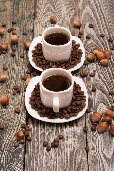 Two small white cups of coffee with cocoa beans, slices of chocolate and hazelnuts on wooden background — Stock Photo, Image