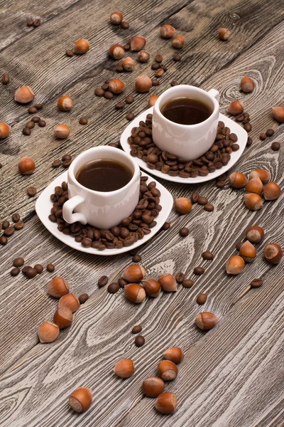 Dos pequeñas tazas de café blanco con granos de cacao, rodajas de chocolate y avellanas sobre fondo de madera — Foto de Stock