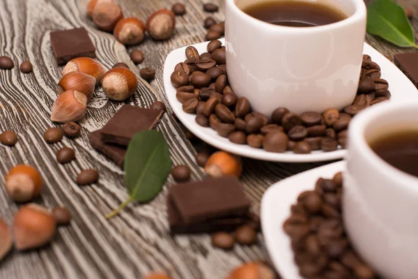 Zwei kleine weiße Tassen Kaffee mit Kakaobohnen, Schokoladenscheiben, Haselnüssen und grünen Blättern auf Holzgrund — Stockfoto