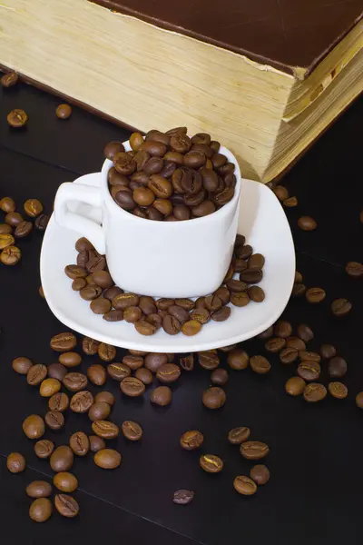 Small white cup with coffee beans and old book on a dark wooden background — Stock Photo, Image