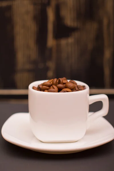Grãos de café em uma xícara branca em um fundo de madeira escura — Fotografia de Stock