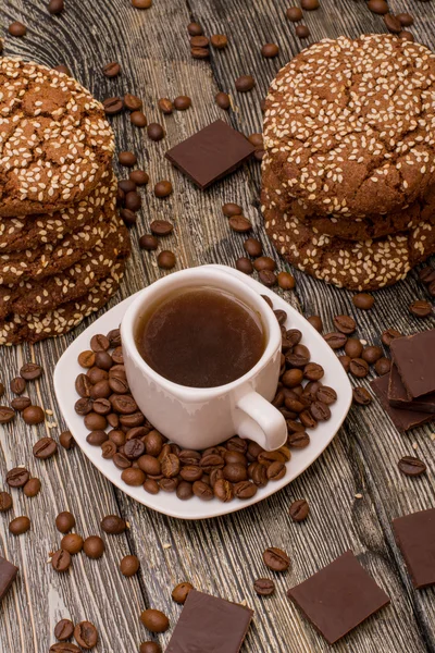 Pequena xícara branca de café, grãos de café torrados, biscoitos com sementes de gergelim, fatias de chocolate no fundo de madeira — Fotografia de Stock