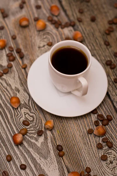 Small white cup of coffee, hazelnuts and cocoa beans on wooden background — Stock Photo, Image