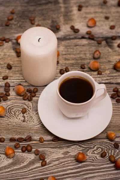 Malý bílý šálek kávy, lískové oříšky, kakaové boby, svíčka na dřevěné pozadí — Stock fotografie