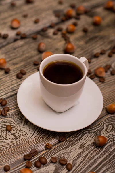 Small white cup of coffee, hazelnuts and cocoa beans on wooden background — Stock Photo, Image