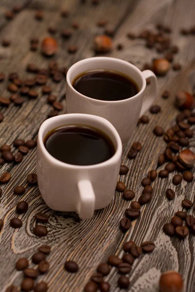 Due piccole tazze bianche di caffè, nocciole e fagioli di cacao su sfondo di legno — Foto Stock