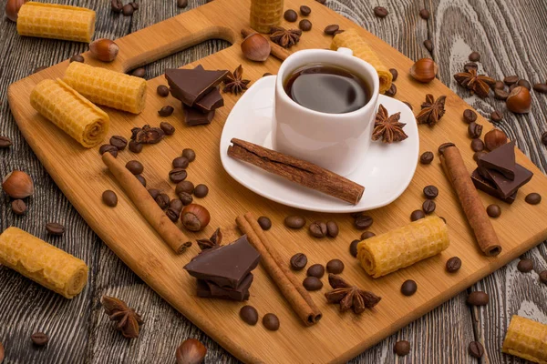 Small white cup of coffee, cinnamon sticks, cocoa beans, star anise, hazelnuts, chocolate, and cookies on wooden background — Stock Photo, Image