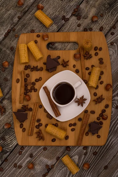 Small white cup of coffee, cinnamon sticks, cocoa beans, star anise, hazelnuts, chocolate, and cookies on wooden background — Stock Photo, Image