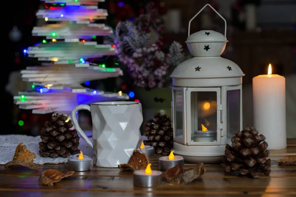 Weihnachtsdekoration mit Lampe mit Kerze, weißer Becher, trockene Zitrusfrüchte, Kegel auf Holztisch vor Lichterhintergrund Stockbild