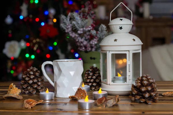 Weihnachtsdekoration mit Lampe mit Kerze, weißer Becher, trockene Zitrusfrüchte, Kegel auf Holztisch vor Lichterhintergrund Stockbild
