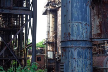 Terk edilmiş bir endüstriyel kompleksin içinde büyüyen yeşillik, boya ve paslı borular, yatay görünüm