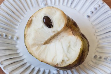 Bir Cherimoya meyvesinin yarısına yakından bakmak Annona Cherimola 'nın beyaz flüt plaka üzerinde merkezi gövde ve karakteristik siyah tohum izlenimini ortaya çıkarır.