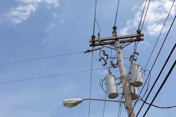 Top of a power pole with multiple transformers, insulators, wires, and a street light against a blue sky with clouds, creative copy space, horizontal aspect