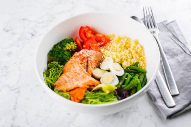 Sağlıklı yemek, keto yemek konsepti. Balık salatası kasesi mermer masa arkasında. Somonlu salata, kuskus, sebze, bıldırcın yumurtası. Yakın plan.