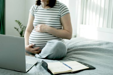 Beautiful pregnant young woman working on a laptop sitting on the bed in the bedroom. Home office, home education. Social distance during quarantine, self-isolation concept. clipart
