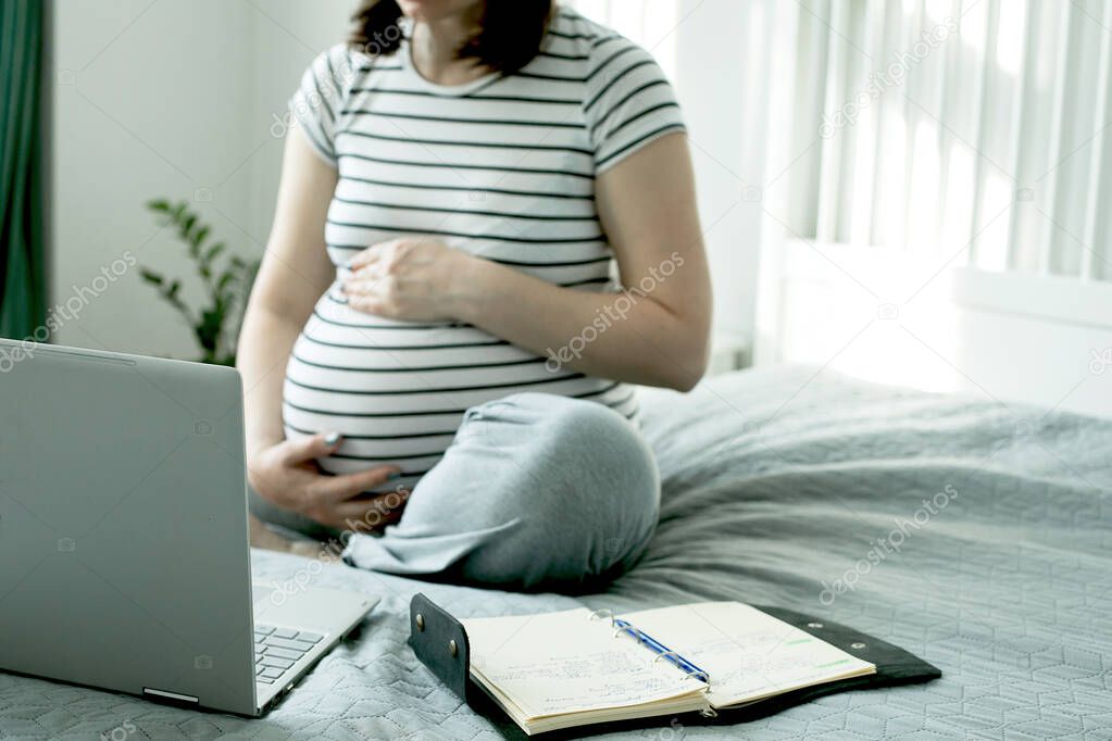 Beautiful pregnant young woman working on a laptop sitting on the bed in the bedroom. Home office, home education. Social distance during quarantine, self-isolation concept.